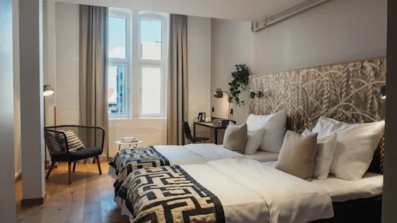 Viihtyisä hotellihuone, jossa kaksi sänkyä, mukava tuoli, työpöytä ja suuri ikkuna.