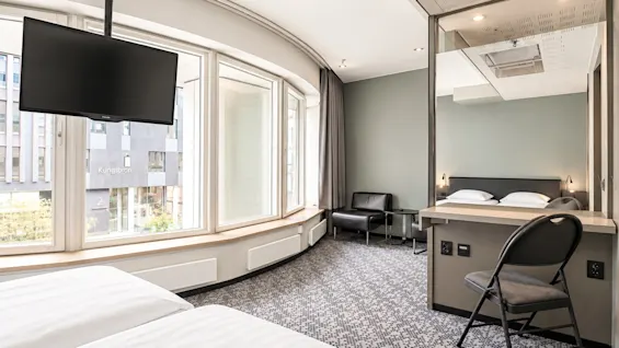 Bright hotel room with large windows, a bed in the foreground, a TV, and a desk with a mirror.