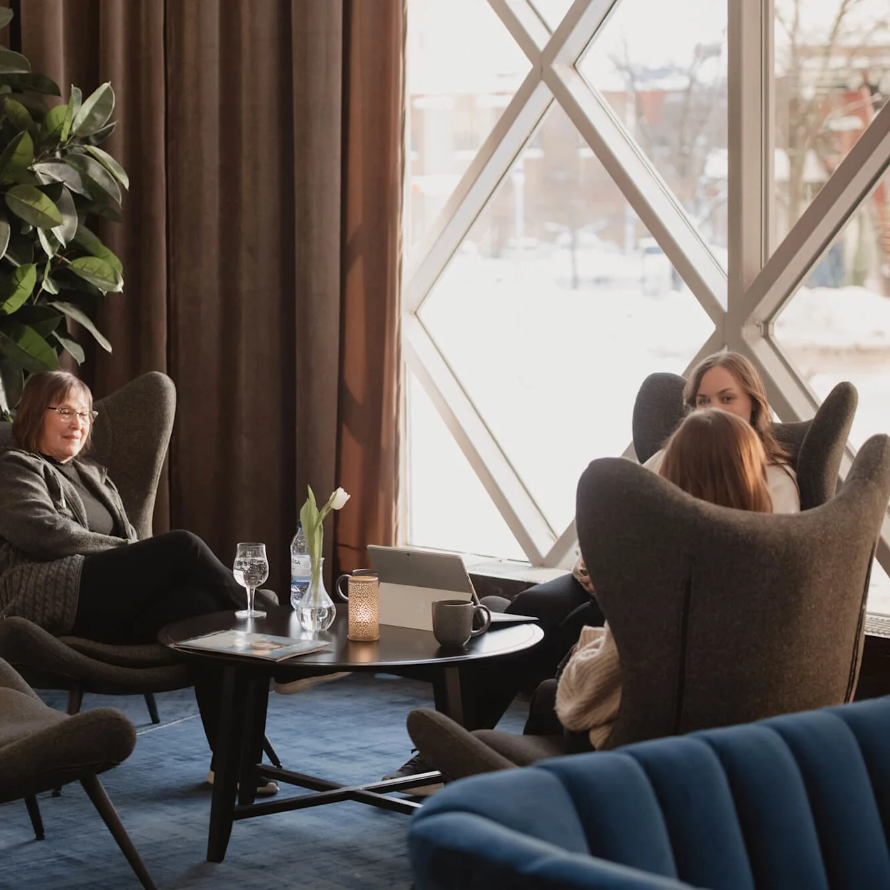A woman sits in a cozy chair, conversing with two others nearby, surrounded by indoor plants and large windows; natural light streams into the lounge area.