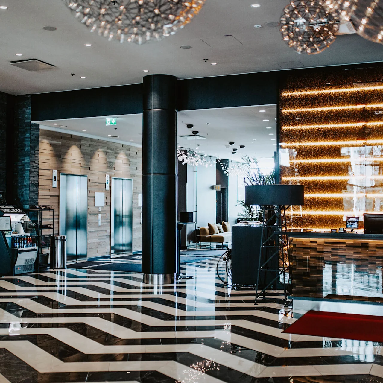 A hotel lobby features shiny, patterned floors and a reception desk lit by warm, horizontal lights. To the left, there are elevators and a display with drinks. "CLAR" is visible on the wall.