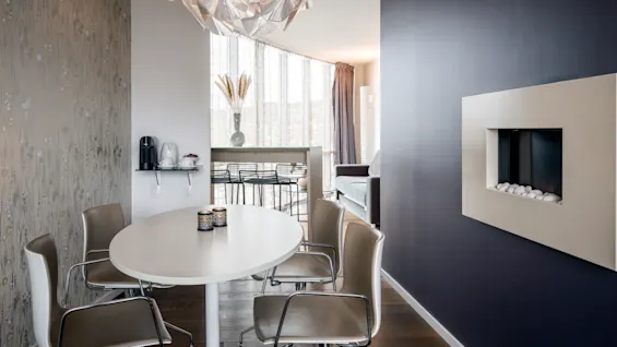 Round dining table with four chairs in a modern room, under an abstract chandelier; a small shelf holds a coffee maker and cups, adjacent to a wall-mounted fireplace.