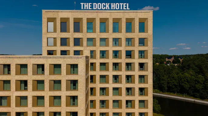 The Dock Hotel, en moderne bygning med mange vinduer, står mod en blå himmel med en grøn skovklædt baggrund.