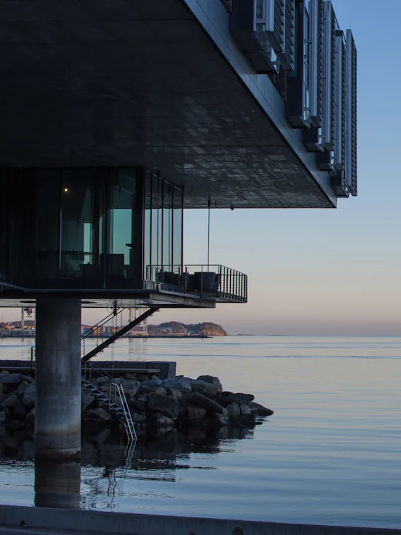 Modern building overhanging calm sea at dusk, pilings and rocks visible, tranquil ocean horizon.
