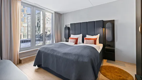 A double bed with dark upholstery and orange cushions in a modern hotel room. Sunlight streams through large windows, revealing a balcony with chairs outside.