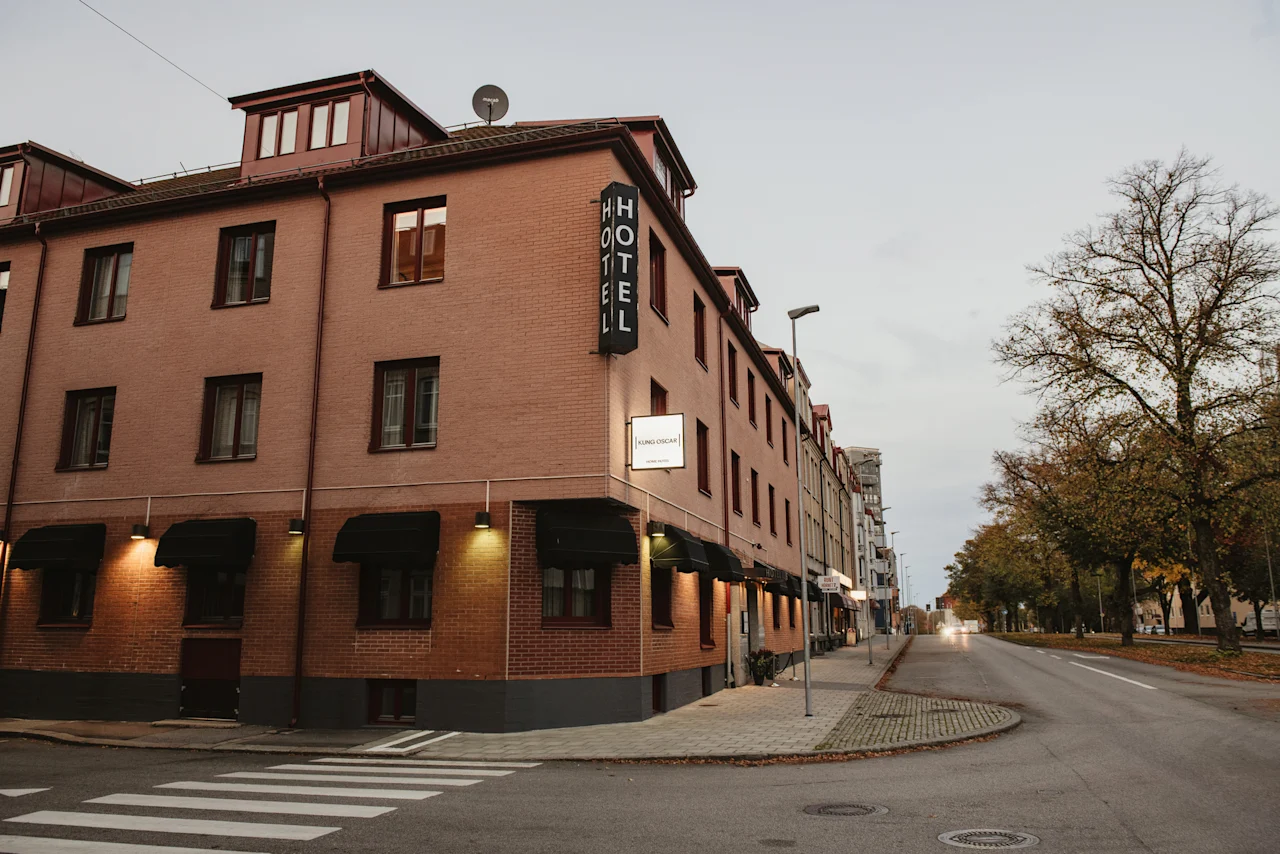 A brick hotel building on a street corner with a HOTEL sign, Home Hotel Kung Oscar. Trees line the street under an overcast sky.