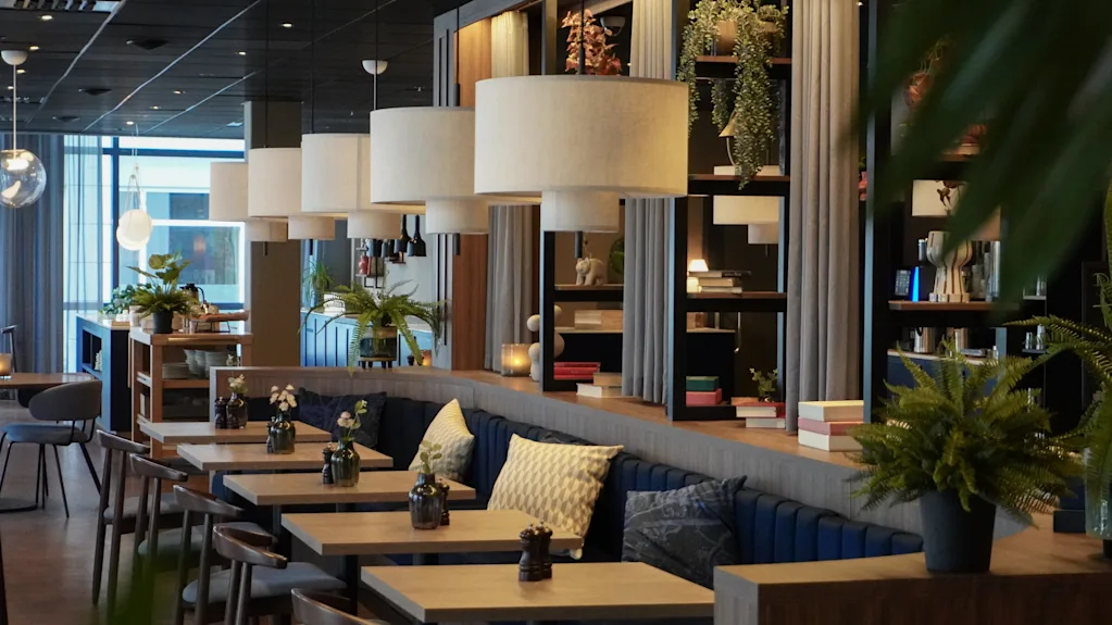 Tables line a stylish, modern cafe; hanging lamps illuminate potted plants and books on wooden shelves, creating a cozy ambiance with cushioned seating.