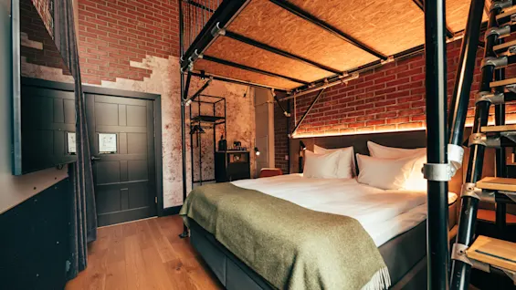A cozy hotel room with an industrial design, featuring a bed with white linens, exposed brick walls, and a loft area.