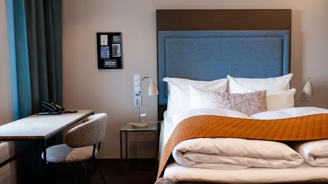 Huolellisesti pedattu sänky, jossa on valkoiset lakanat ja oranssi huopa, sijaitsee modernissa hotellihuoneessa. Vasemmalla on pieni pöytä ja tuoli ikkunan alla.