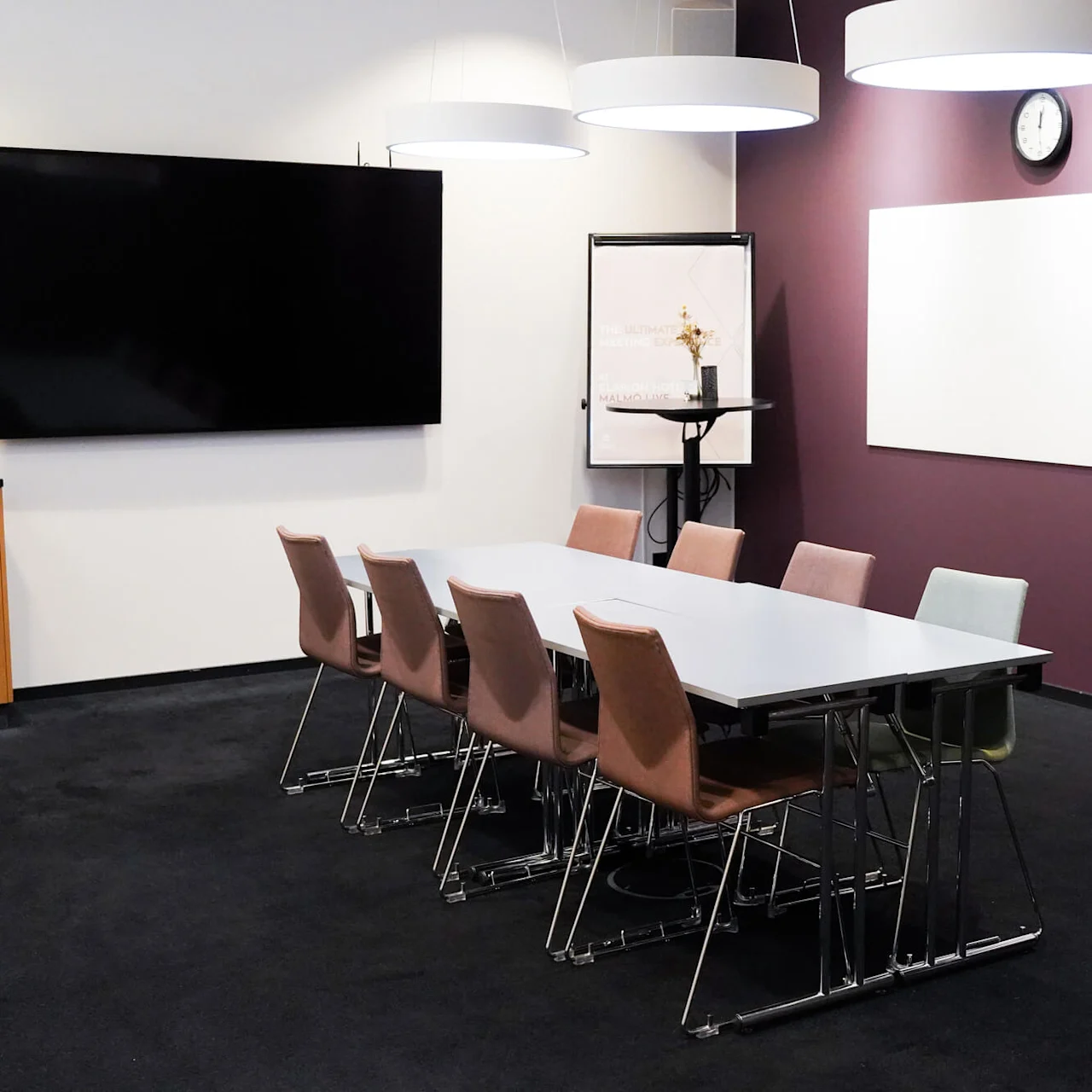 Et møterom har et bord med åtte stoler, omgitt av lilla vegger. En stor skjerm og klokke henger på veggene. Et flippover-stativ står i hjørnet.