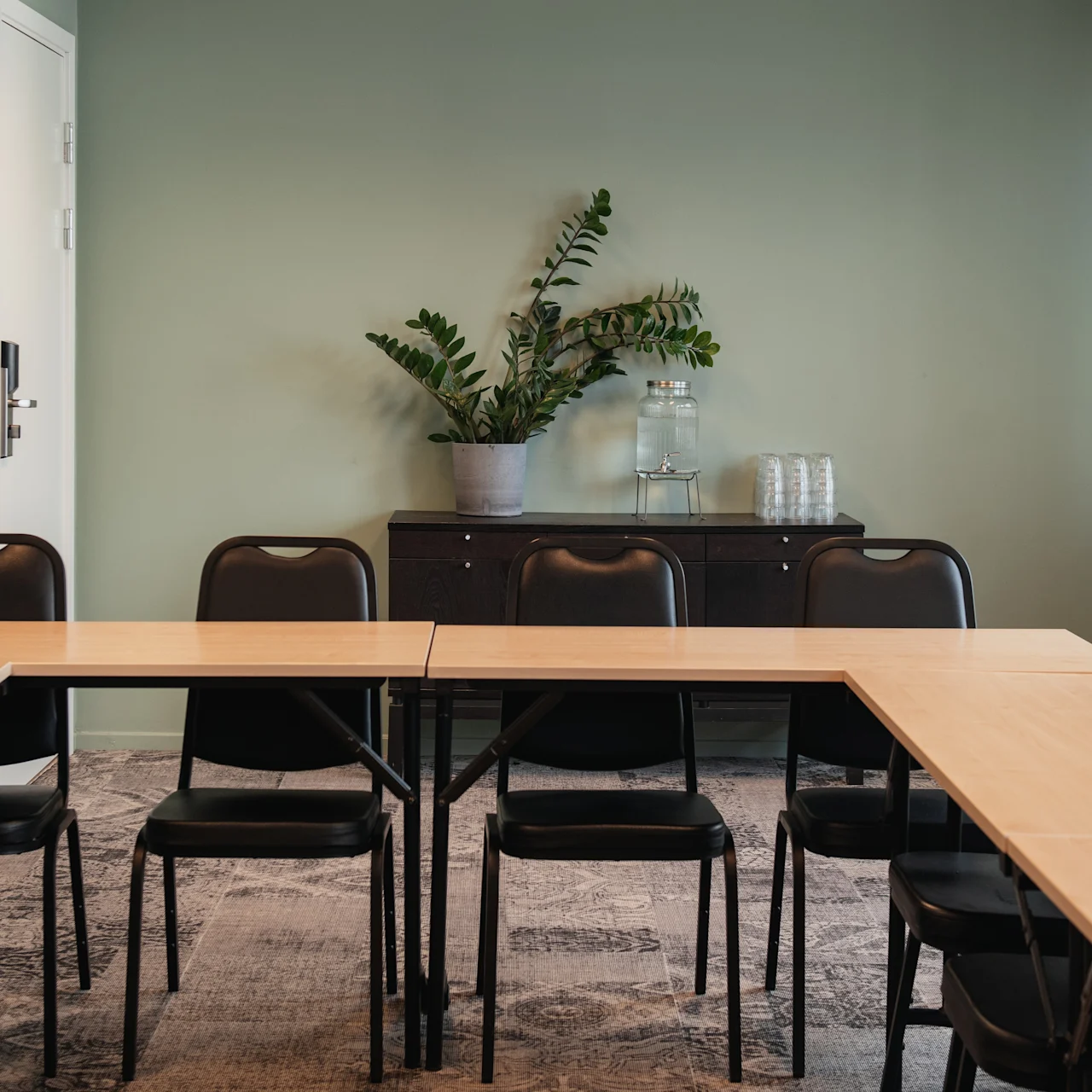 Tables form a U-shape in a meeting room with black chairs; a cabinet with a plant and water glasses is against a green wall.