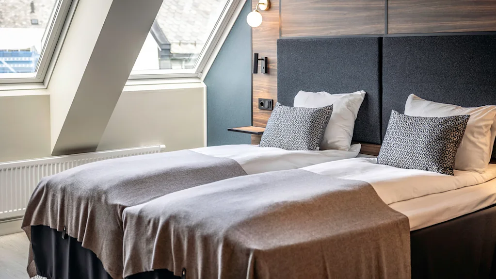 To pent oppredde enkeltsenger med grå tepper og mønstrede puter står i et moderne rom. Store vinduer slipper inn naturlig lys, med en panelvegg og lampe over sengegavlen.