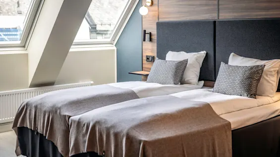 Två prydligt bäddade enkelsängar med gråa filtar och mönstrade kuddar står i ett modernt rum. Stora fönster ger naturligt ljus, med en träpanel och lampa ovanför sängen.