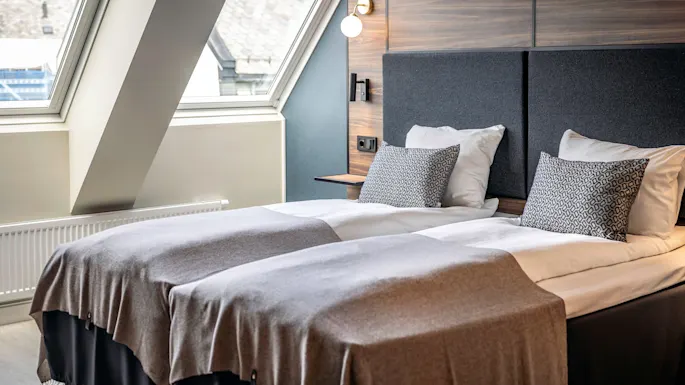 Two neatly made twin beds with grey blankets and patterned cushions sit in a modern room. Large windows provide natural light, with a wood-paneled wall and lamp above the headboard.