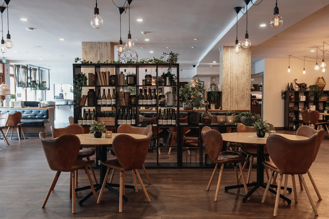 A cozy restaurant interior with wooden tables, leather chairs, and shelves filled with bottles and books, illuminated by warm hanging lights.