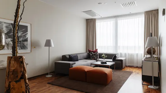 Ett mysigt och modernt vardagsrum med en grå L-formad soffa, orange puffar och ett stort fönster med skira gardiner.