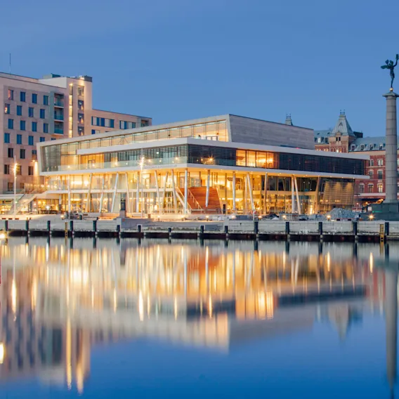 En modern byggnad med stora fönster avger varmt ljus, stående vid en vattenkant. Historiska tegelbyggnader och en staty är upplysta i bakgrunden och speglas i lugnt vatten.