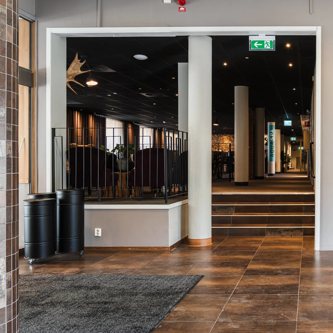 Hissdörrar stängda, bredvid en svart hylla med pappersmaterial; i en kaklad hotellobby med skyltar "WC" och klocka ovanför. Skyltar visar: "AQUA SPA," pilar och WC-riktningar.