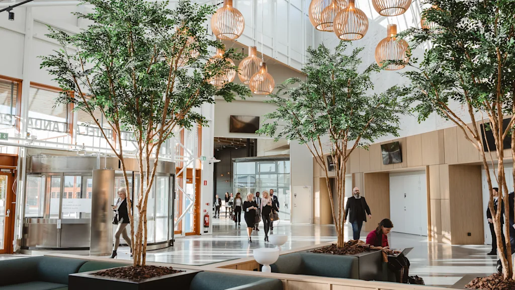 A bright, modern hotel lobby with people moving and relaxing. Indoor trees and unique lighting create a welcoming atmosphere. Arlanda.
