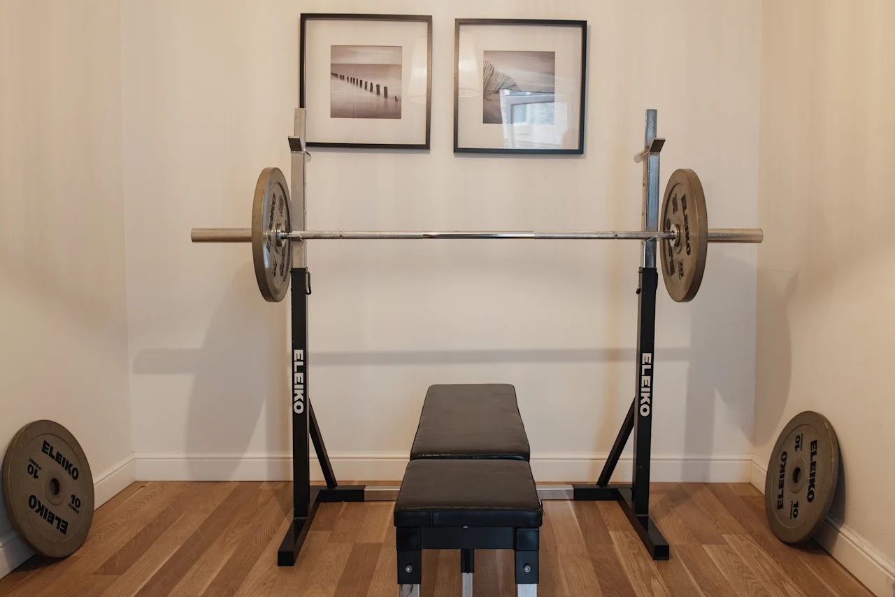 Et vægtløftningsudstyr i et rum, med en bænk, vægtstang med vægte og et stativ. To indrammede billeder pryder væggen.