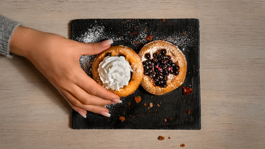 A hand is taking a pastry with whipped cream from a slate board with two pastries, on a wooden table.