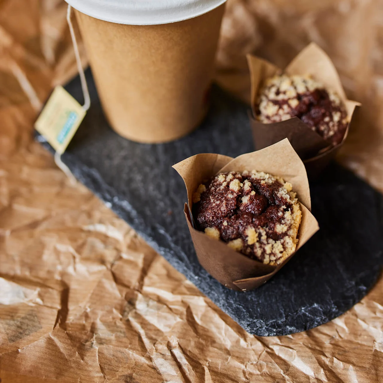 En brun papirkopp med hvitt lokk står på krøllete papir ved siden av to smuletopperte sjokolademuffins på en mørk skiferplate. En tepose-streng henger fra koppen.