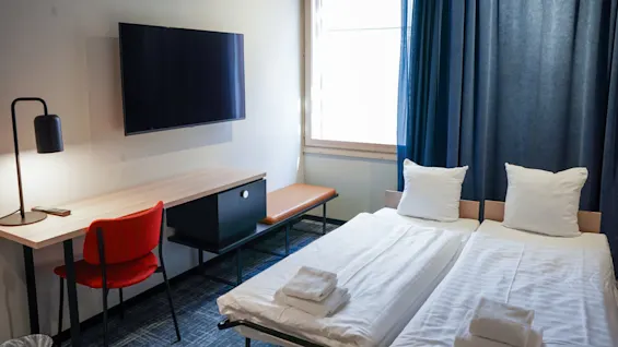 Siisti hotellihuone, jossa on vierekkäin kaksi sänkyä, seinälle kiinnitetty televisio kirjoituspöydän yläpuolella, punainen tuoli ja ikkuna sinisillä verhoilla.