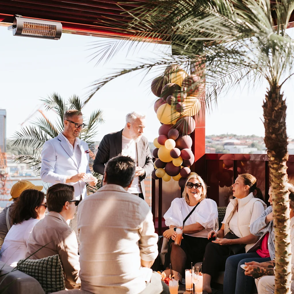 People socializing and enjoying drinks on a sunny rooftop terrace in Göteborg, with palm trees and balloons in the background.