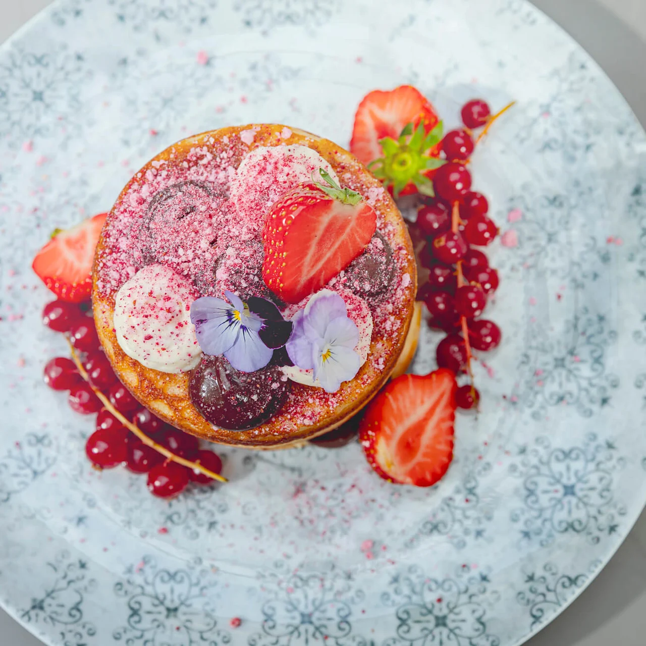 A stack of pancakes, topped with strawberries, cream, decorative flowers, and powdered sugar, sits on a decorative plate with berry garnish, providing an elegant presentation.