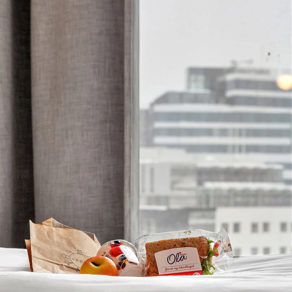 A sandwich, an apple, and yogurt rest on a white bed with a cityscape visible through a window. Text on packaging: "Brødrene Ola fersk og håndlaget".