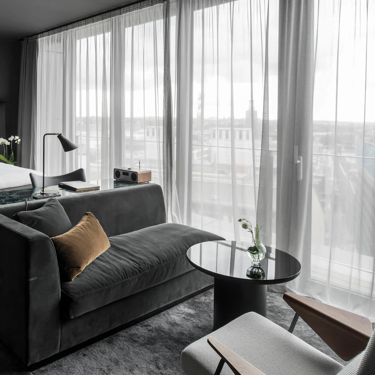 Et ryddig hotellrom har en stor seng, en mørk sofa og et glassbord med blomster, innrammet av gulv-til-tak-vinduer med tynne gardiner.