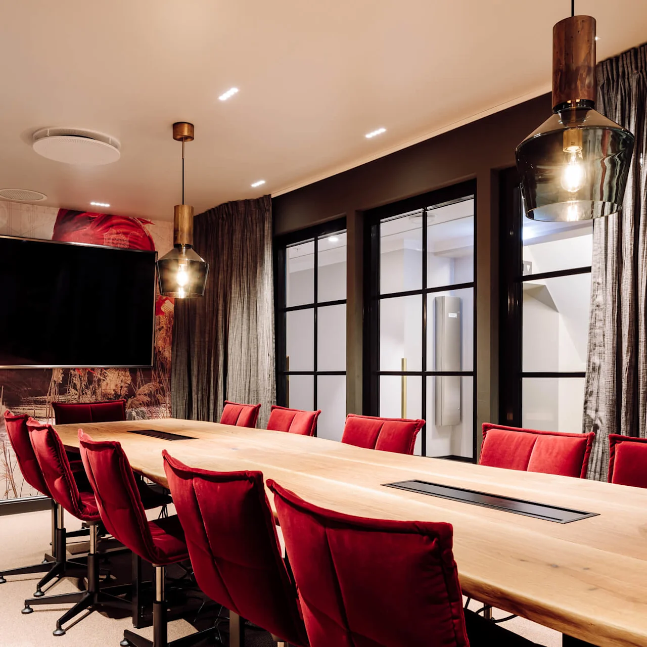 A long wooden conference table surrounded by red chairs, under modern pendant lights. A large screen and artistic wall decor provide a stylish setting.