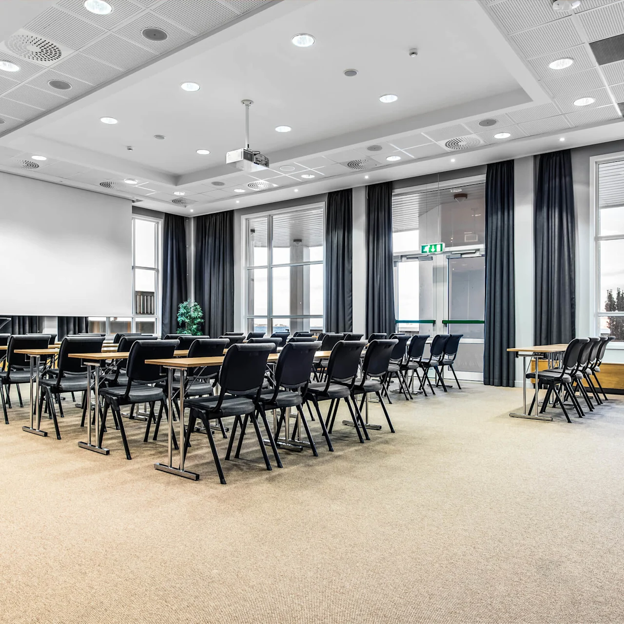 Konferencelokale med rækker af tomme sorte stole og træborde vendt mod en stor hvid skærm. Godt oplyst med loftlys, vinduer med gardiner og et synligt "EXIT"-skilt.