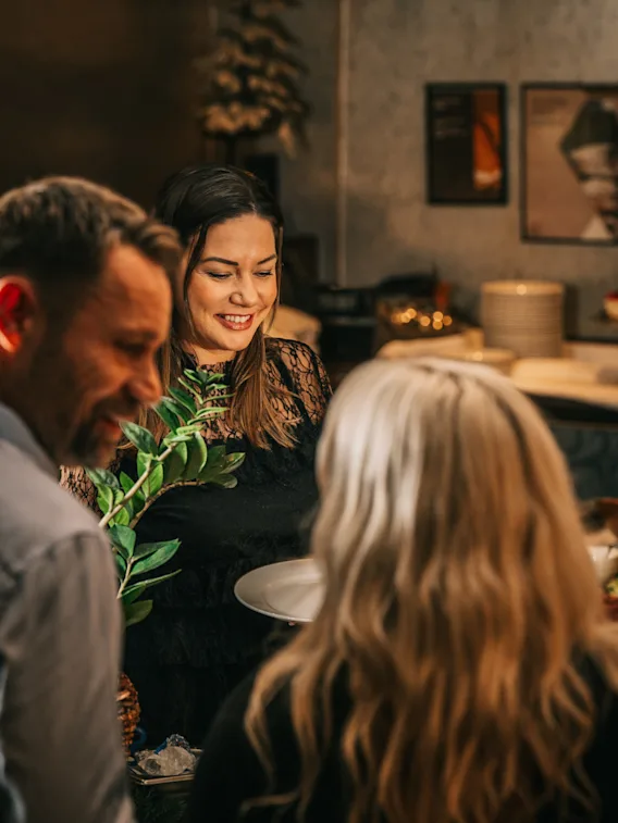 A smiling woman serving food to guests at a cozy restaurant buffet.