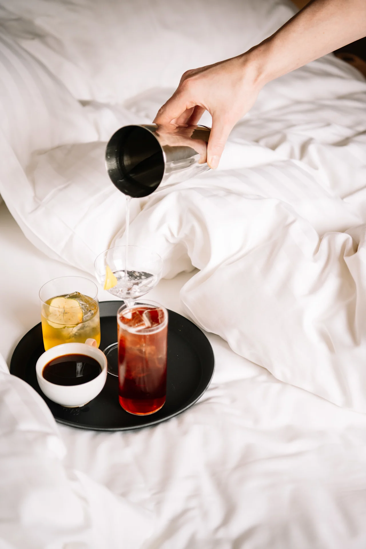 Käsi kaataa cocktailia ravistimesta lasiin valkoisella sängyllä olevalla tarjottimella.