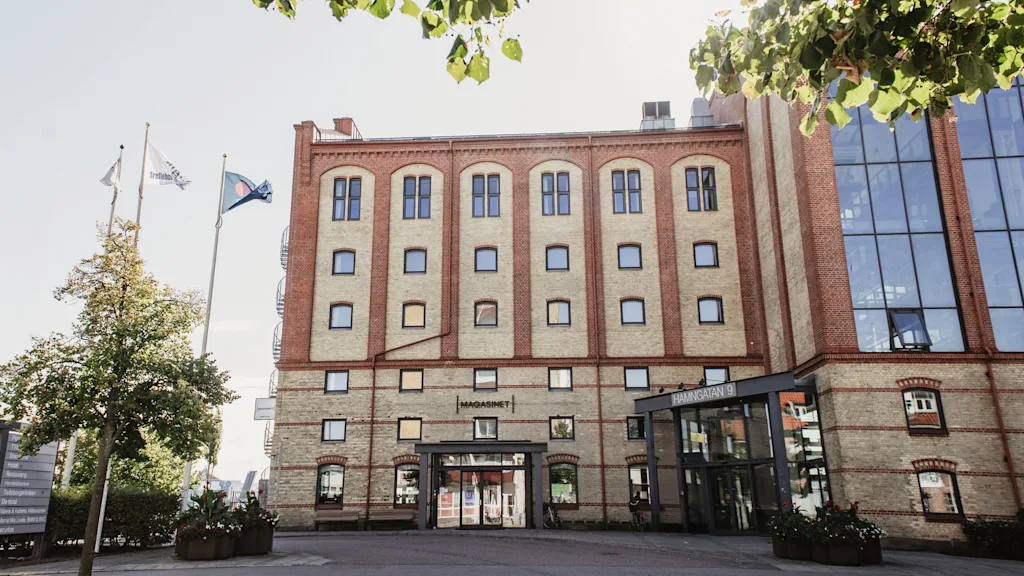 Suuri tiilirakennus, jossa modernit lasiosat ja sisäänkäynti. Liput ja puut kehystävät aurinkoista näkymää. MAGASINET, HAMNGATAN 9.