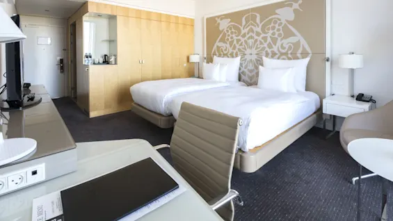 Ett hotellrum innehåller två fint bäddade sängar med vita lakan. En dekorativ huvudgavel, träskåp och ett skrivbord med stol skapar en lugn, modern miljö.
