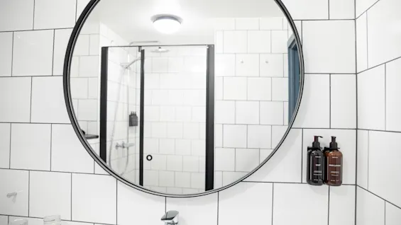 Et rundt spejl reflekterer et flisebelagt badeværelse med en brusekabine af glas. To vægmonterede sæbedispensere er ved siden af spejlet. En vask med hane og gennemsigtige kopper er nedenunder.
