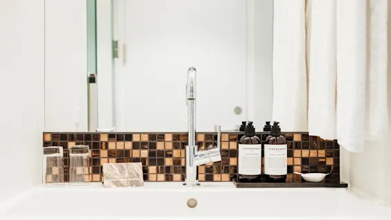 En moderne vandhane i et badeværelse omgivet af mosaikfliser. To glas, lotion og håndsåbe mærket "SPREKENHUS" er pænt arrangeret. Hvide håndklæder hænger i nærheden.