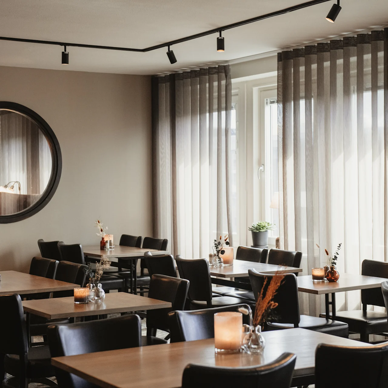 Tables and chairs are neatly arranged in a cozy, softly-lit restaurant, decorated with lit candles and small vases. A large round mirror hangs on the wall, reflecting ambient light.