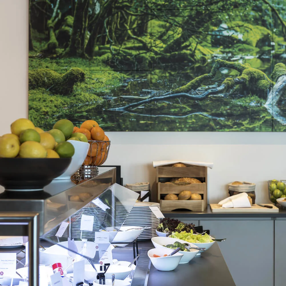 A buffet features bowls of fresh fruit and salads on a counter, with bread and cereals in jars behind. A nature-themed artwork hangs on the wall above.