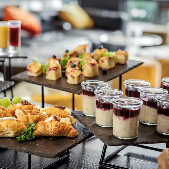Et utvalg av appetittvekkere, inkludert croissanter, smørbrød, fargerike desserter i glasskrukker og fruktspyd, er elegant utstilt på en svart skifer i en lys, uformell spiseinnstilling.