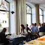 A dining area with people eating at tables near large windows; a salad bar in the foreground offers various ingredients. Sunlight illuminates the modern, bright space.