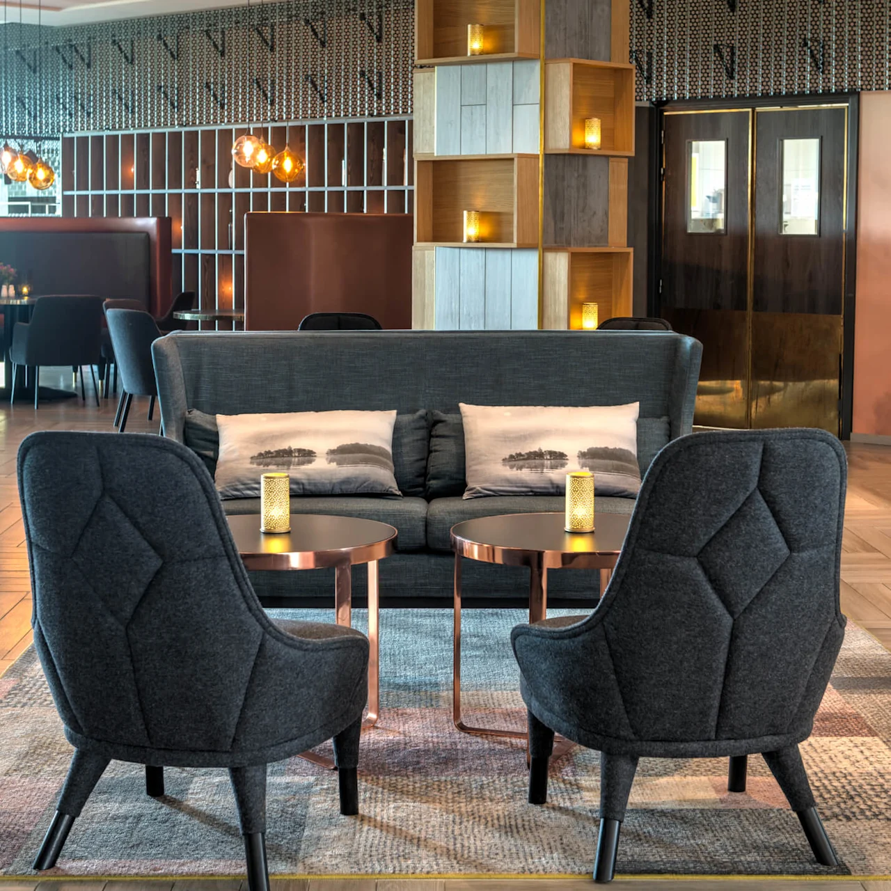 A seating area features two dark chairs and a sofa with patterned pillows, surrounding round tables with lit candles, in a stylish, modern room with decorative shelving and warm lighting.
