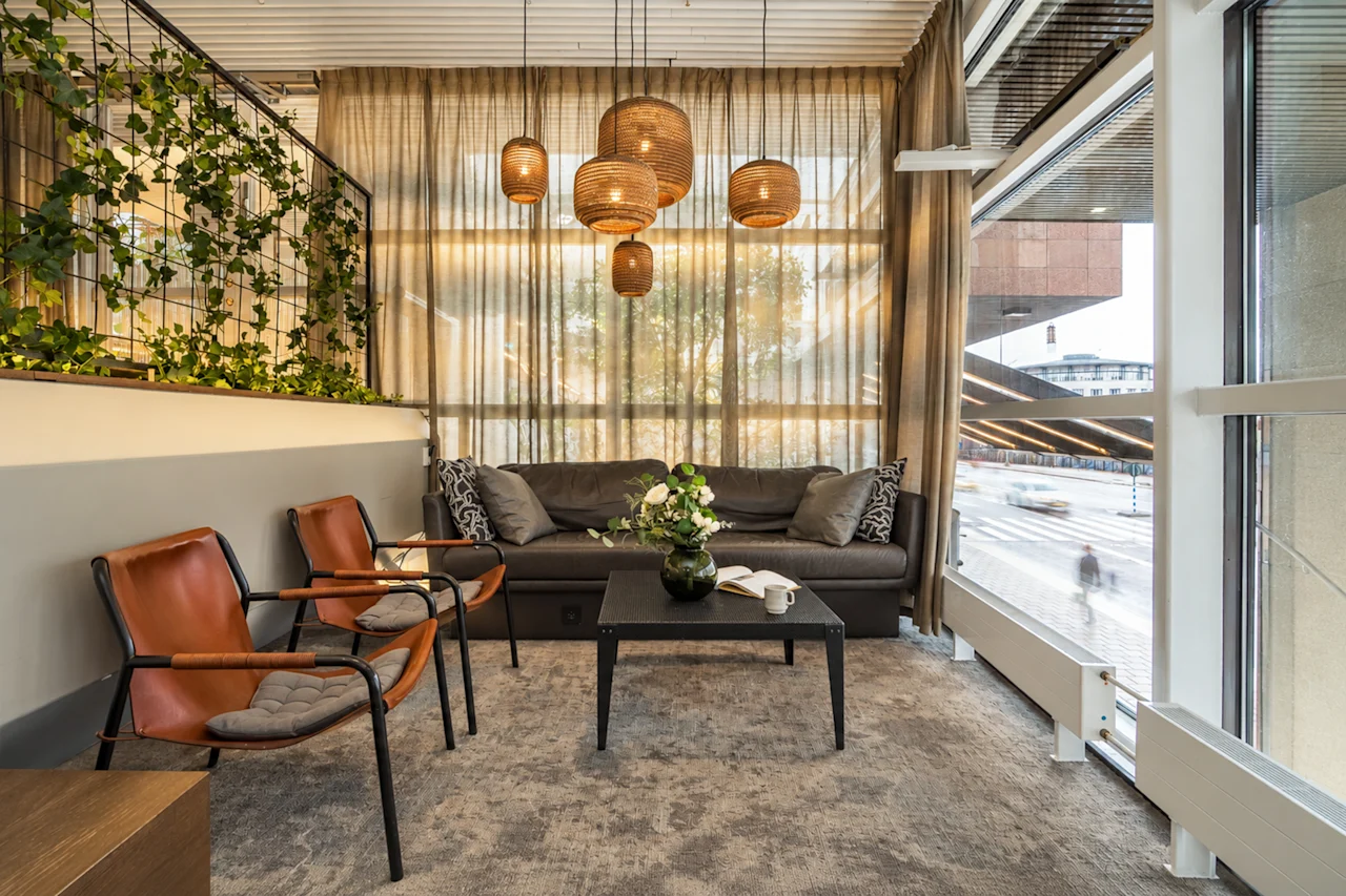 Cozy hotel lobby lounge with a dark sofa, brown leather armchairs, a coffee table, and hanging wicker lamps by a window.