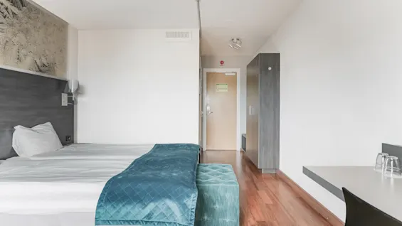 A bright hotel room with a bed, a desk, and a wardrobe. A green exit sign is visible above the door.