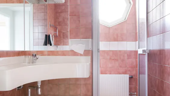 A white sink with a glass and faucet is mounted on a wall with pink tiles in a bathroom, featuring a mirrored cabinet and a shower area with an octagonal window.