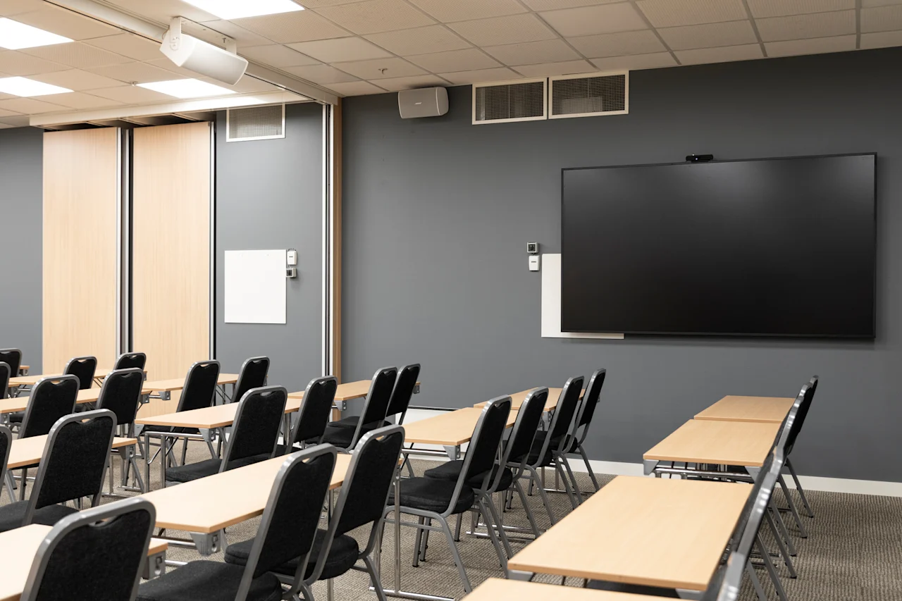 A modern conference room with rows of tables and chairs facing a large display screen on a dark wall, ready for a presentation or meeting.