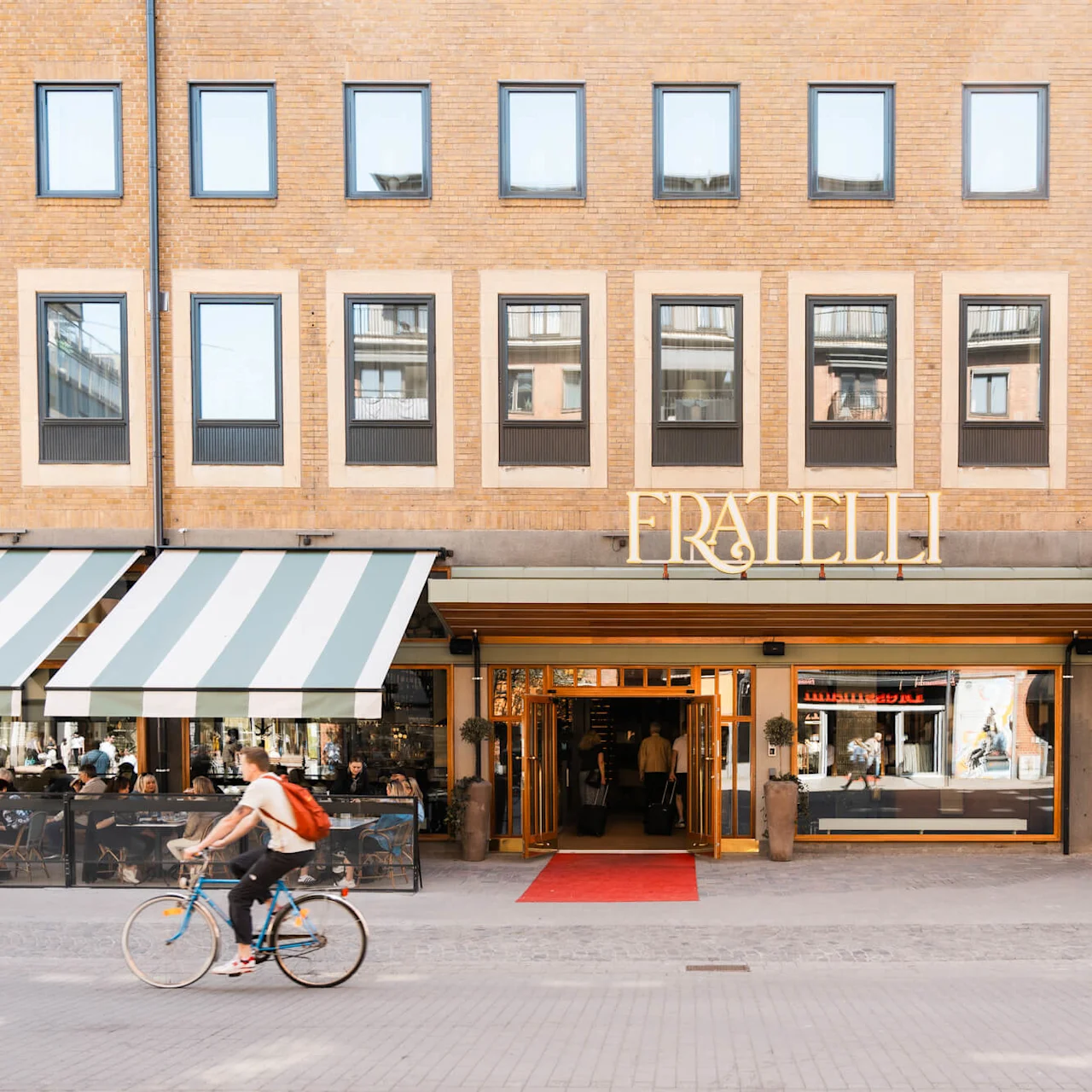 A cyclist rides past the entrance of a building with a sign "FRATELLI" above, large windows, and outdoor seating under striped awnings on a sunny day.