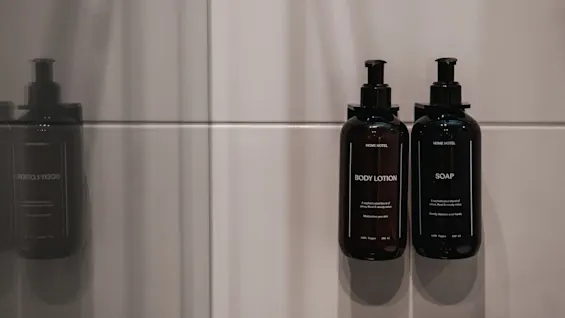 To mørke flasker med body lotion og såpe på en flislagt vegg. Etiketter: BODY LOTION, Fukter huden din. SOAP, Renser skånsomt hendene dine.