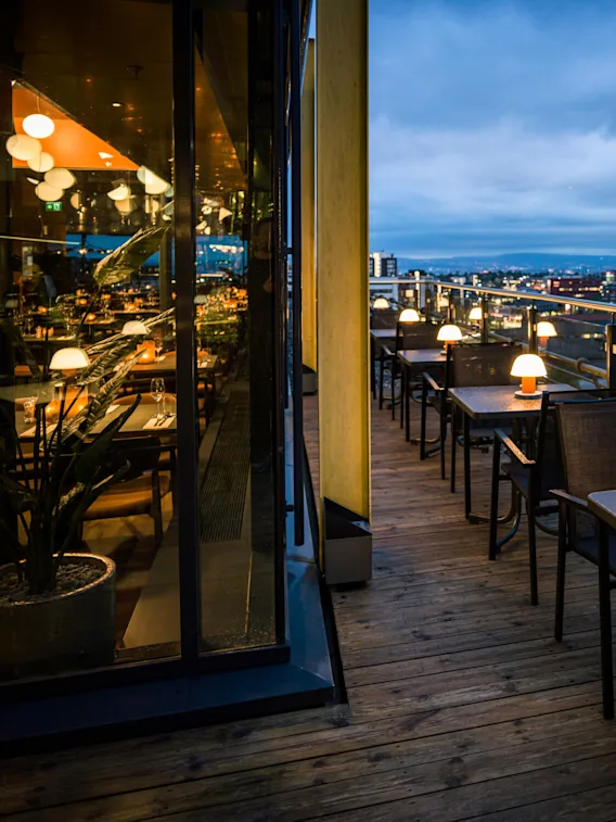 Outdoor dining tables with glowing lamps line a wooden terrace. The cityscape, visible at dusk beyond a glass railing, features a lit building displaying the word "TILIT."